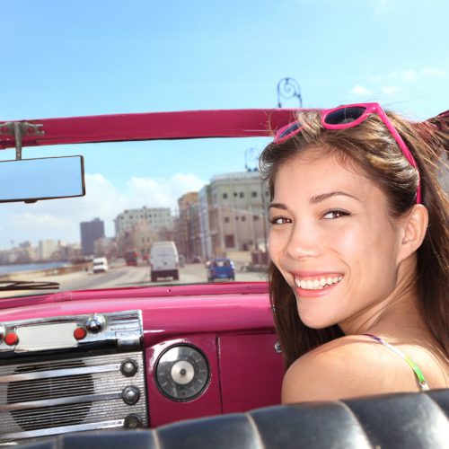 Car woman smiling happy on passenger seat in pink vintage convertible car. Young mixed race Asian / Caucasian female model during cuba vacation. Driving on Malecon waterfront, Havana, Cuba